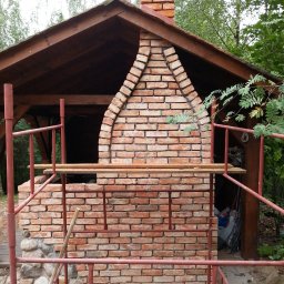 KTM-BUD JACEK LUBACZEWSKI - Murowana konstrukcja z cegły z łukowym wejściem i kominem, częściowo osłonięta drewnianym dachem, otoczona rusztowaniem.