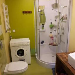 KTM-BUD JACEK LUBACZEWSKI - Wnętrze łazienki z kabiną prysznicową, pralką i toaletą, na ścianach żółto-zielona farba, widoczny drewniany blat umywalki