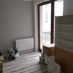 Pok&oacute;j po remoncie: szare ściany, grzejnik, panele podłogowe imitujące drewno, okno balkonowe, białe p&oacute;łki i szafki, kartony i puszki z farbą.