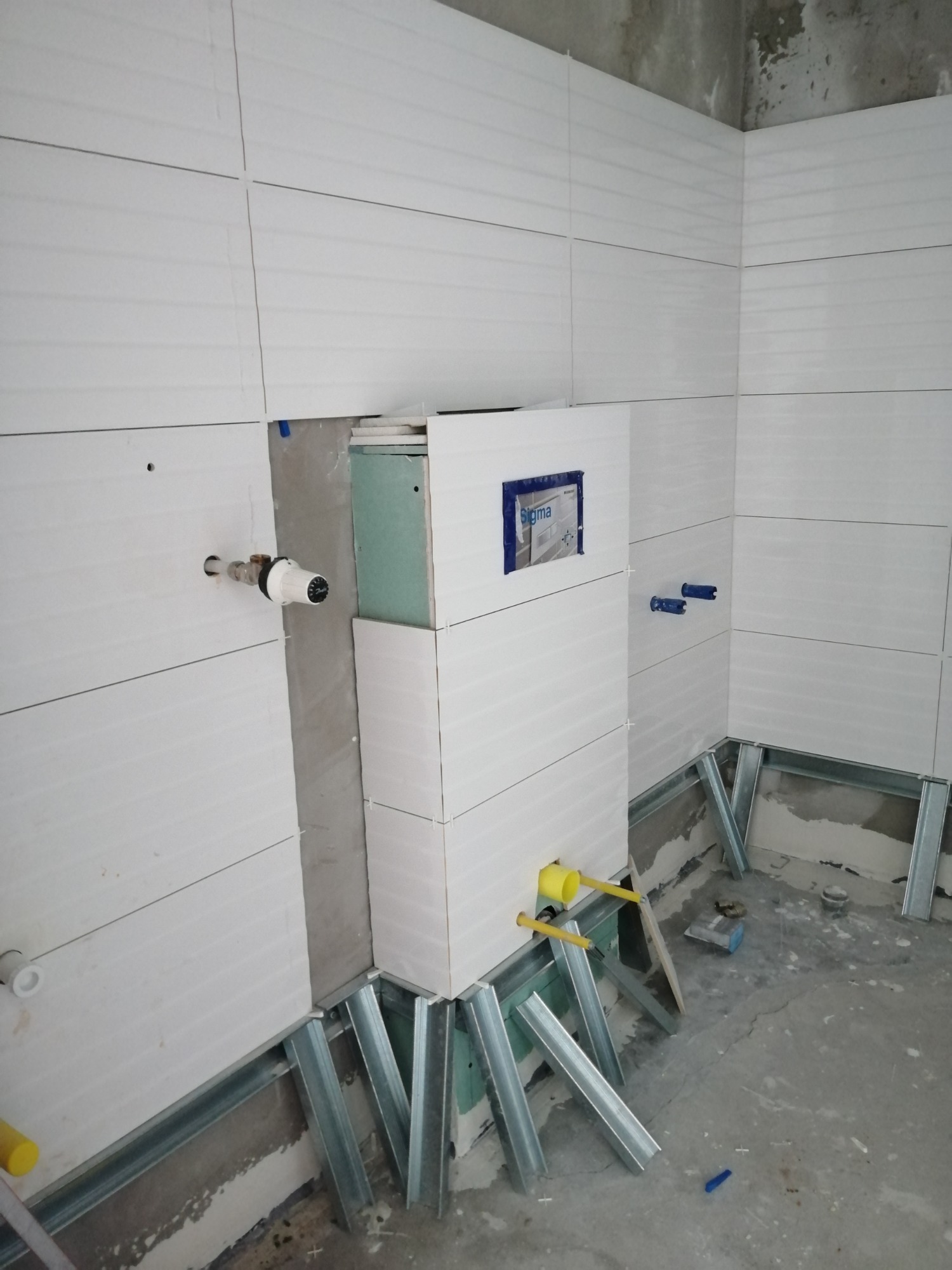 Białe, poziome płytki ścienne z delikatną fakturą w trakcie montażu w łazience, widoczne metalowe profile konstrukcyjne pod zabudowę, instalacja wodna z zaworami.