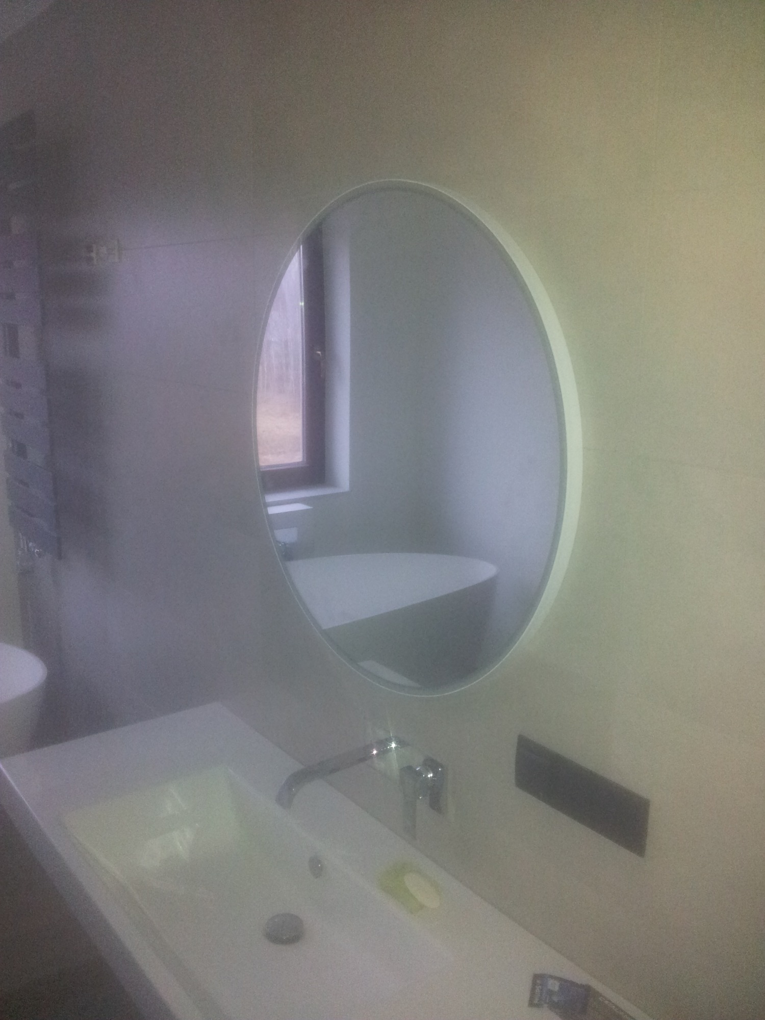 Nowoczesna umywalka z baterią i okrągłym lustrem, w którym odbija się wanna i okno w jasnej łazience wykończonej płytkami.