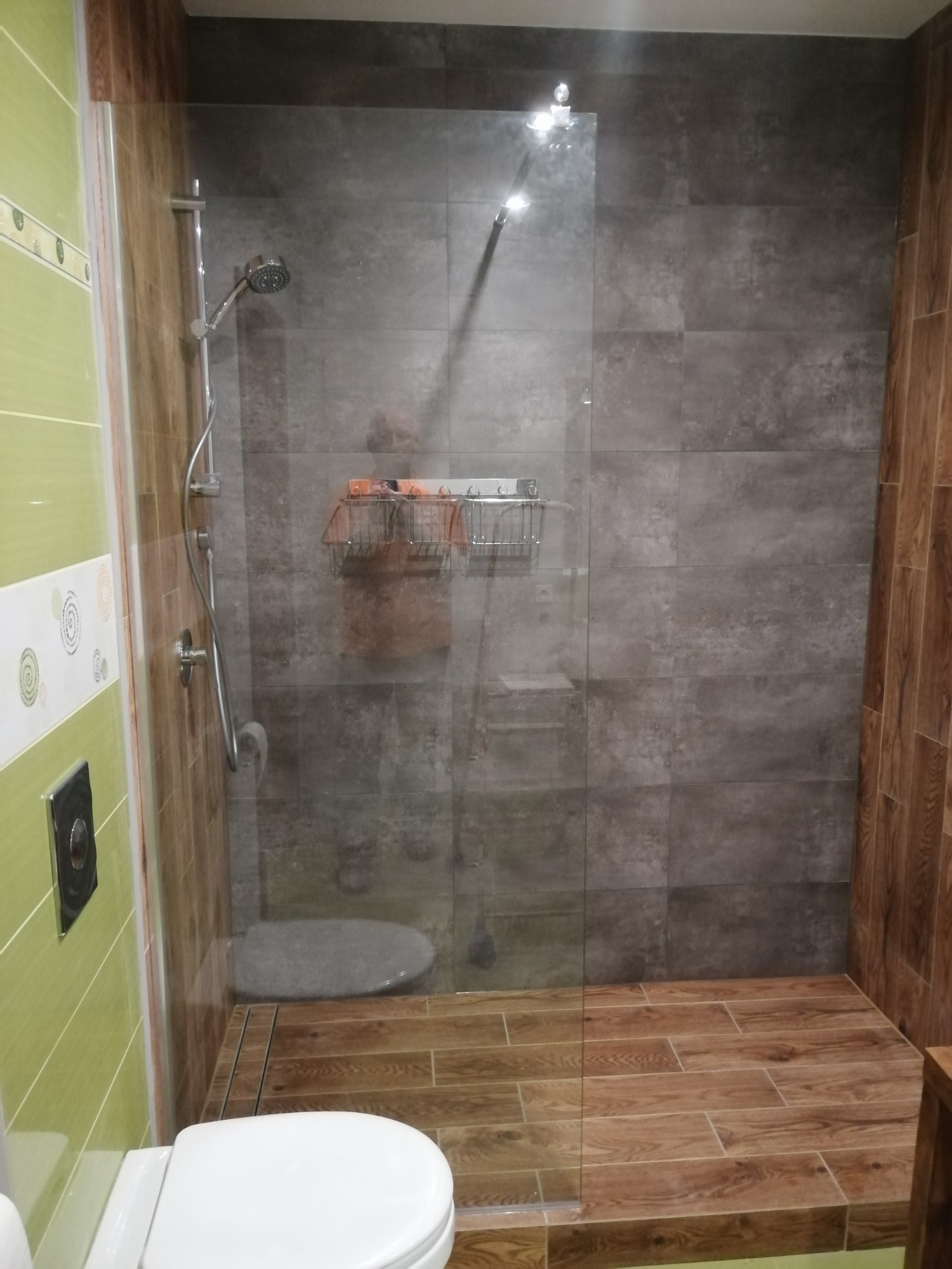 Nowoczesna kabina prysznicowa typu walk-in z szarą glazurą imitującą beton, drewnopodobną podłogą i zielonymi płytkami na ścianie z widoczną toaletą.