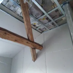 Montaż sufitu podwieszanego z płyty gipsowo-kartonowej z widoczną drewnianą konstrukcją wsporczą i metalowym rusztem, izolacja widoczna w przestrzeni między sufitem a płytami.