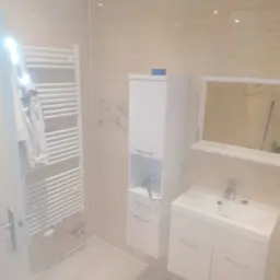 Jasna łazienka z beżowymi płytkami, białymi meblami, grzejnikiem drabinkowym i lustrem, po remoncie.