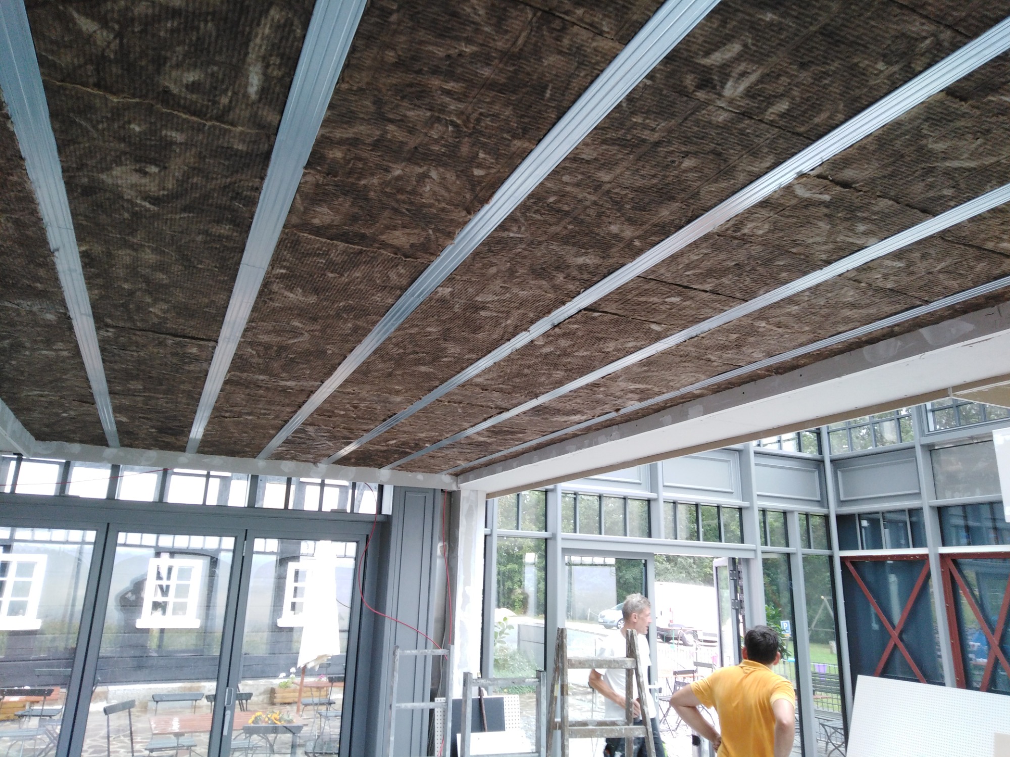 Montaż izolacji z wełny mineralnej na suficie podwieszanym, widoczne metalowe profile konstrukcyjne, pracownicy na drabinie w przeszklonym pomieszczeniu.