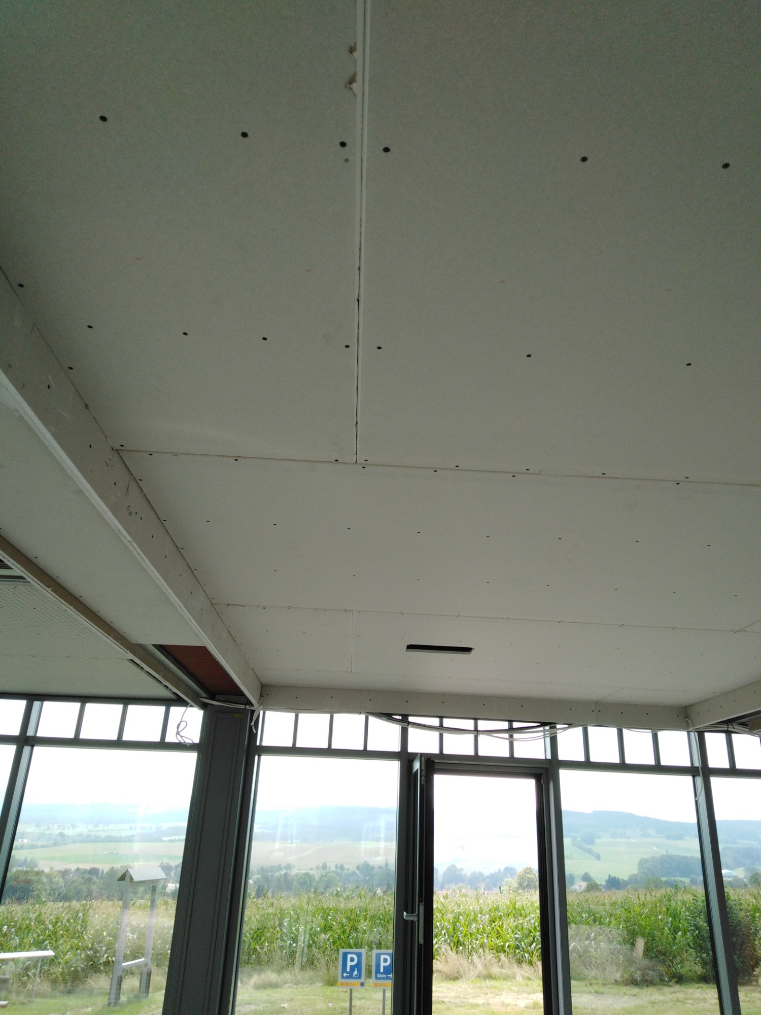 Sufit z płyt kartonowo-gipsowych w trakcie montażu, widoczne ślady po wkrętach, z widokiem na pole kukurydzy przez duże okna.