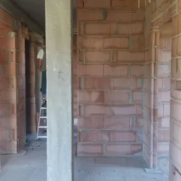 Wnętrze murowanego domu w trakcie budowy, widoczne ściany z czerwonej cegły dziurawki, betonowe słupy, podłoga wylewka, pracownik na drabinie w tle.