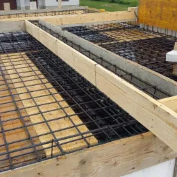 Zbrojenie płyty fundamentowej z drewnianym szalunkiem i elementami podtrzymującymi, widok z góry na placu budowy.