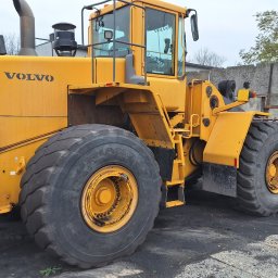 Centrum mechaniki i transportu CEMIT Łukasz Więtczak - Żółta ładowarka Volvo L220E z dużymi oponami i łyżką, stojąca na placu budowy. Widoczny fragment betonowego muru w tle. Sprzęt budowlany.