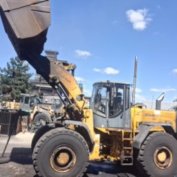 Centrum mechaniki i transportu CEMIT Łukasz Więtczak - Żółta ładowarka kołowa na placu budowy, z podniesioną łyżką i widocznymi elementami konstrukcyjnymi. W tle zabudowania przemysłowe i błękitne niebo.