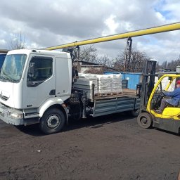 Centrum mechaniki i transportu CEMIT Łukasz Więtczak - Biała ciężarówka z paletą materiałów budowlanych i żółty wózek widłowy z operatorem na błotnistym placu, pod żółtym rurociągiem.