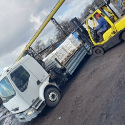 Centrum mechaniki i transportu CEMIT Łukasz Więtczak - Żółty wózek widłowy załadowuje palety z workami na pakę białej ciężarówki na błotnistym placu. Operator w kasku i roboczym ubraniu. W tle instalacja przemysłowa.