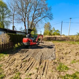 Centrum mechaniki i transportu CEMIT Łukasz Więtczak - Czerwona mini koparka wyrównuje teren podwórka, przygotowując podłoże pod budowę. Widoczne ślady opon i rozplantowany gruz. W tle zieleń i błękitne niebo.