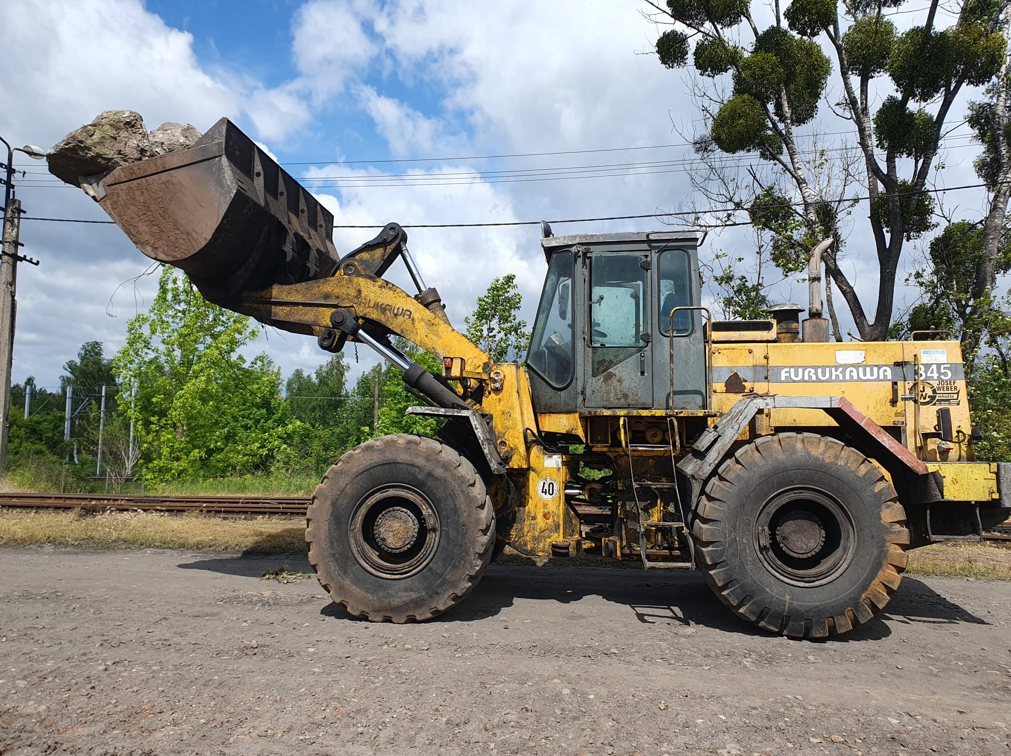 Żółta ładowarka Furukawa 345 z pełną łyżką kamieni, stojąca na żwirowej drodze obok torów kolejowych, z drzewami w tle i błękitnym niebem z chmurami.