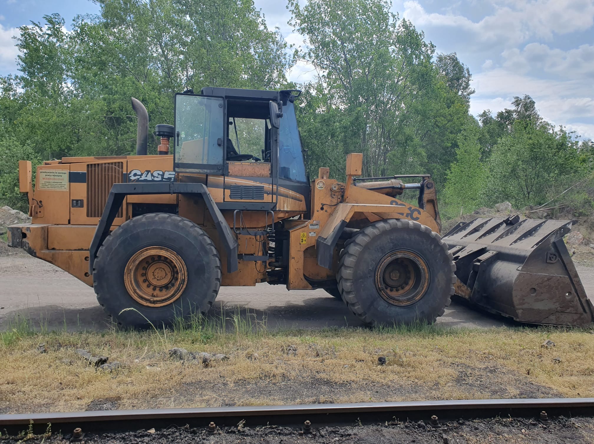 Pomarańczowa ładowarka kołowa CASE na żwirowym podłożu, z widocznym łyżką i elementami konstrukcyjnymi, na tle zieleni i błękitnego nieba z chmurami.