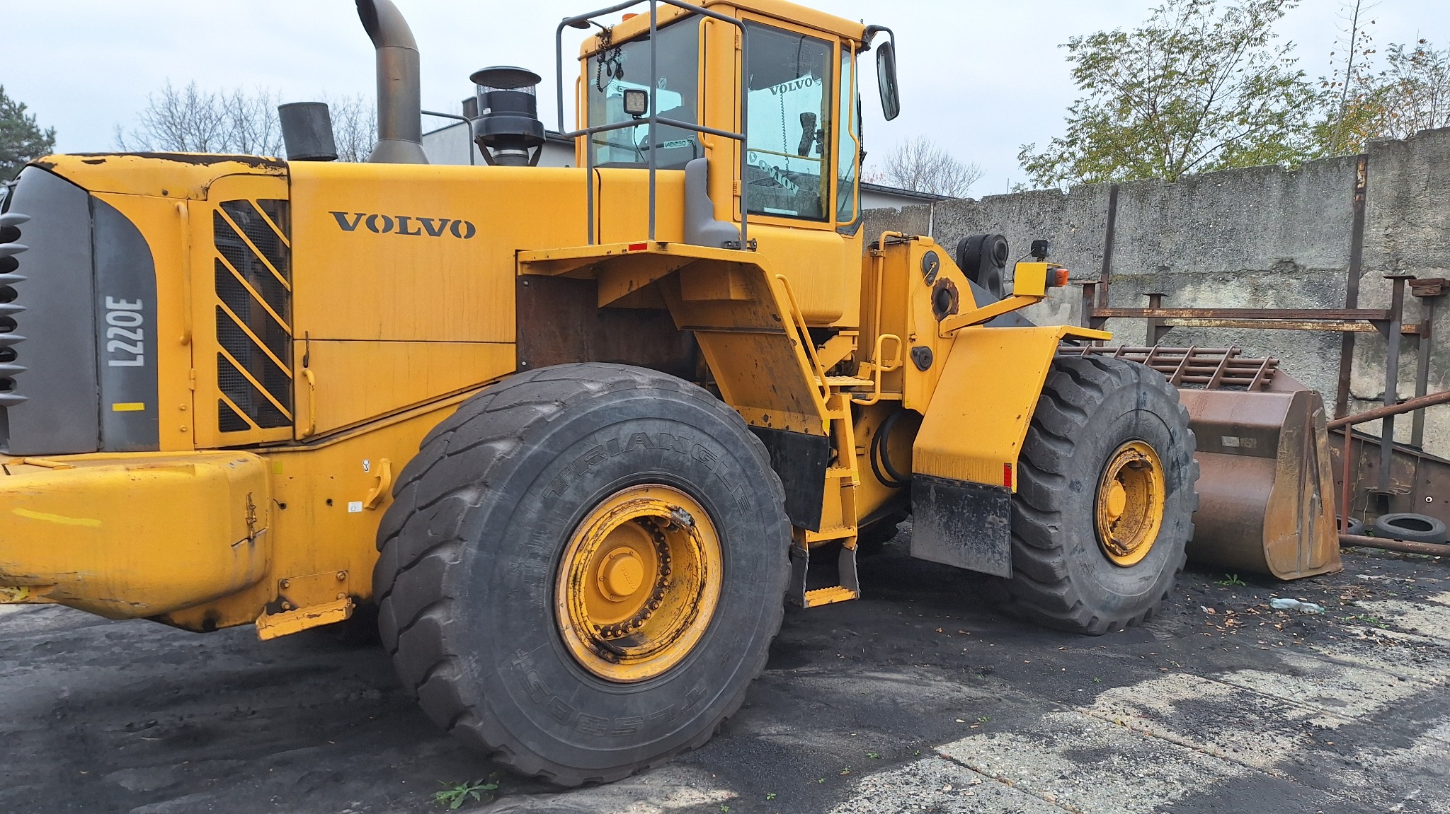 Żółta ładowarka Volvo L220E z dużymi oponami i łyżką, stojąca na placu budowy. Widoczny fragment betonowego muru w tle. Sprzęt budowlany.