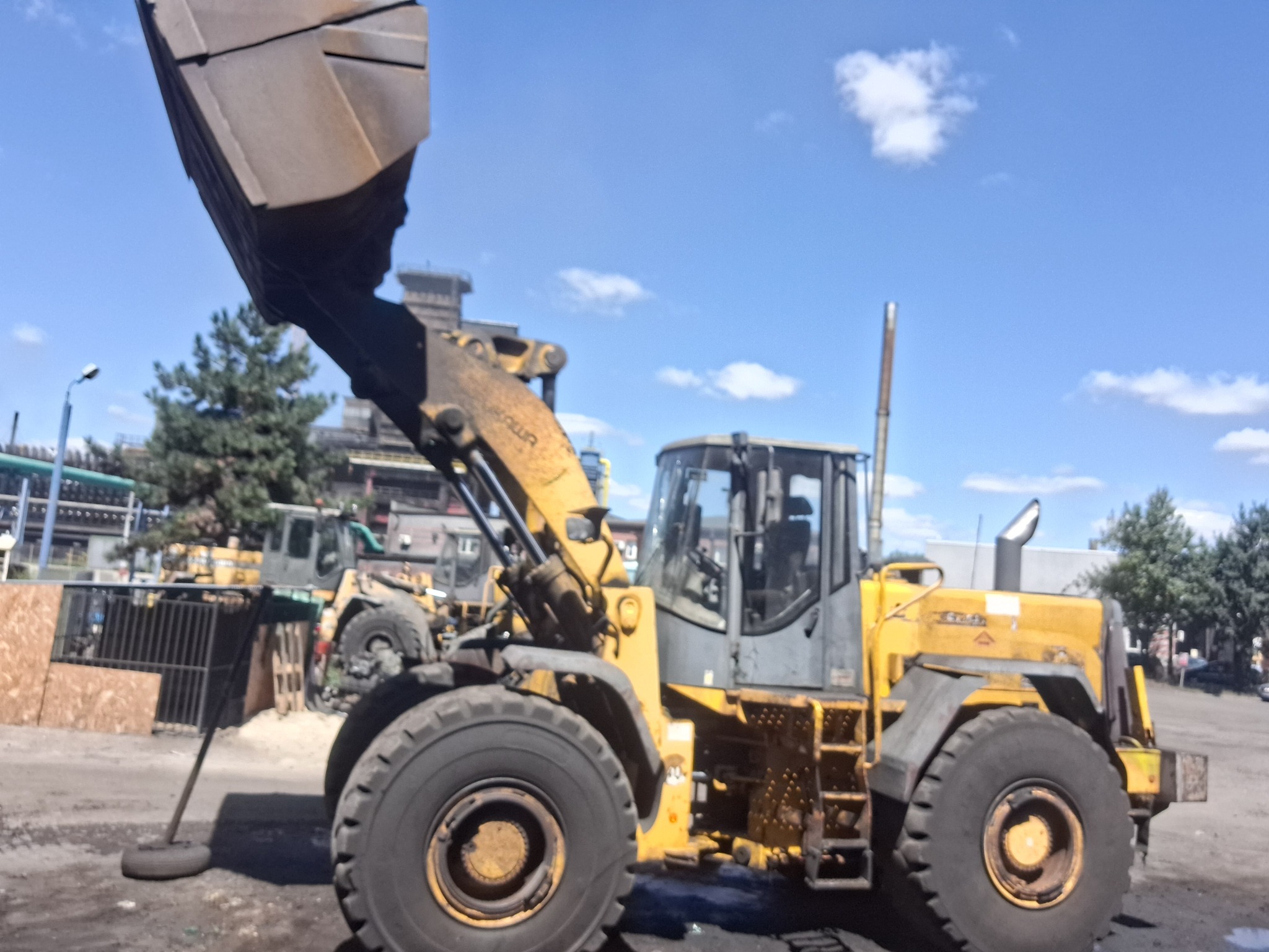 Żółta ładowarka kołowa na placu budowy, z podniesioną łyżką i widocznymi elementami konstrukcyjnymi. W tle zabudowania przemysłowe i błękitne niebo.