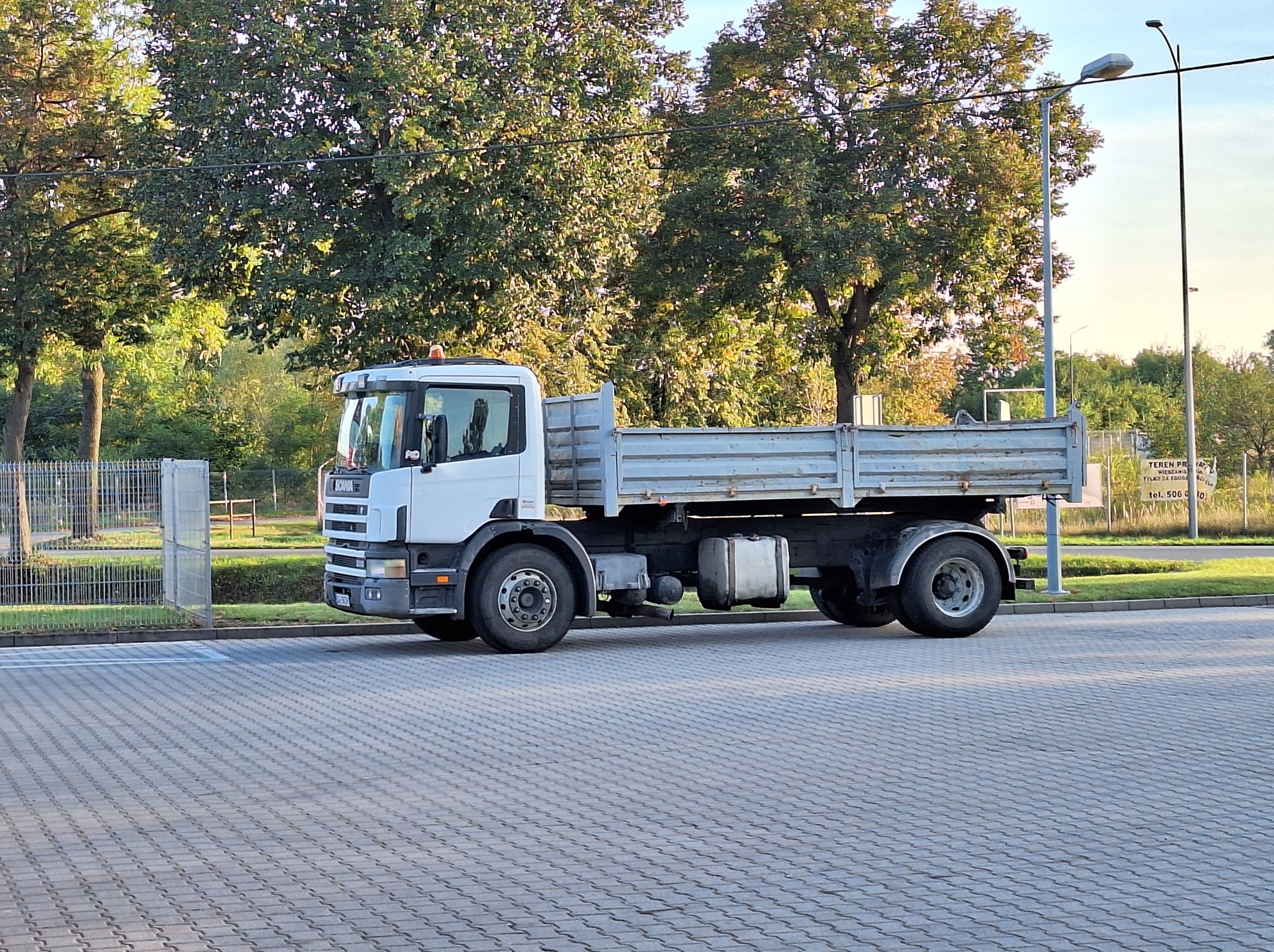 Biała ciężarówka Scania z otwartą skrzynią ładunkową, zaparkowana na brukowanym placu, otoczona zielenią drzew i krzewów w słoneczny dzień.