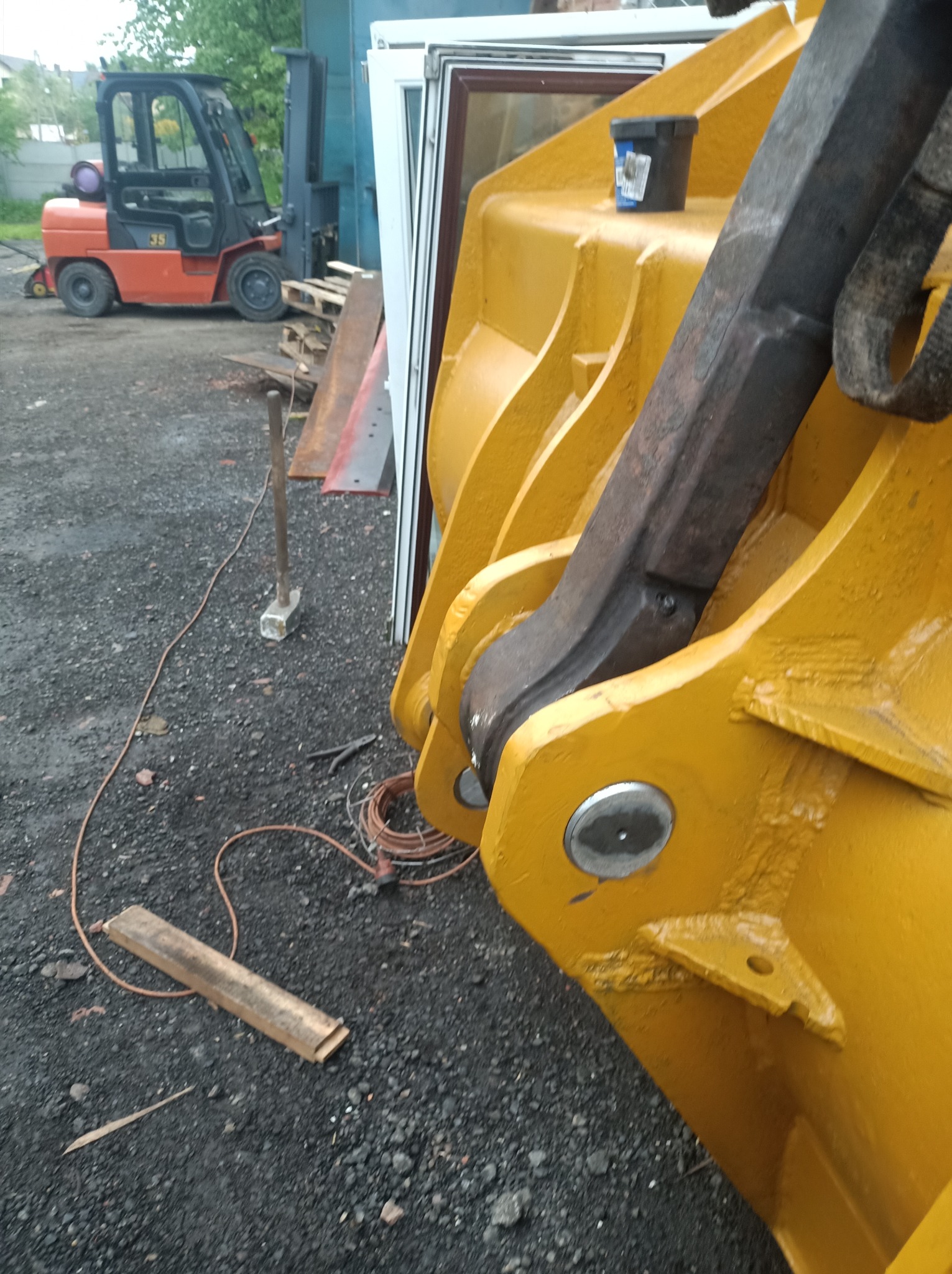 Żółta łyżka koparki z widocznymi spawami i mocowaniami, na tle żwirowego podłoża i wózka widłowego. Detal konstrukcyjny maszyny budowlanej.