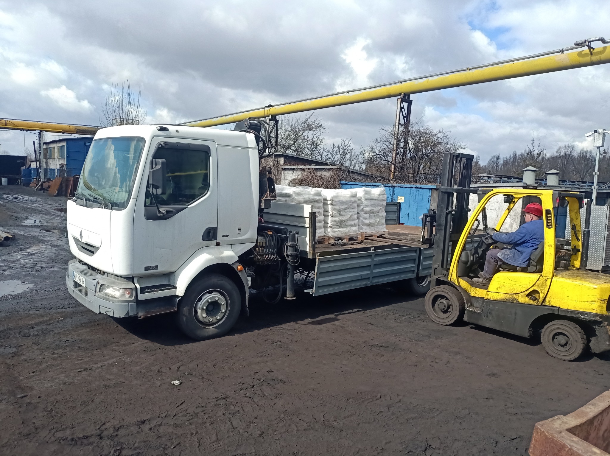 Biała ciężarówka z paletą materiałów budowlanych i żółty wózek widłowy z operatorem na błotnistym placu, pod żółtym rurociągiem.