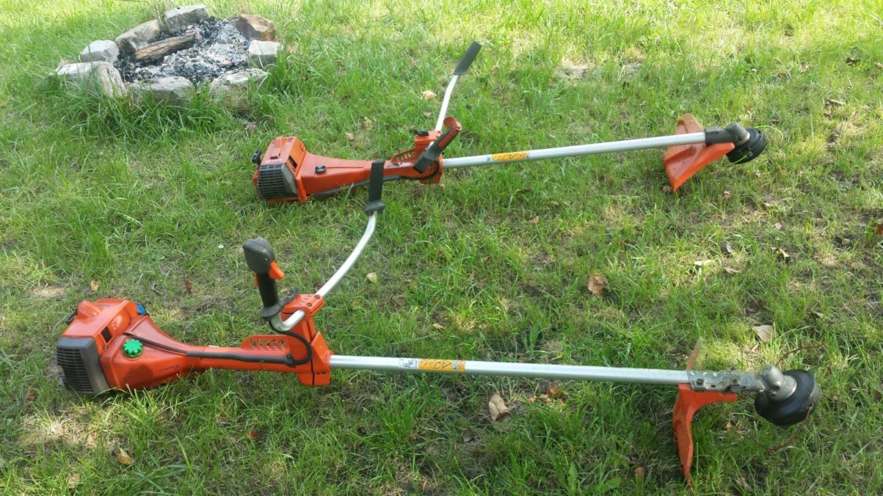 Dwie pomarańczowe podkaszarki spalinowe Husqvarna leżące na trawie, w tle palenisko obłożone kamieniami.