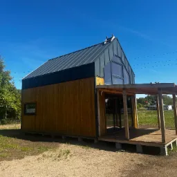 Nowoczesny, drewniany dom modułowy z szarą, metalową dachówką i werandą, osadzony na betonowych blokach, na tle błękitnego nieba i zieleni.