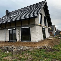 GPF FIRMA HANDLOWO USŁUGOWA - Surowy stan domu jednorodzinnego z czarnymi oknami i roletami zewnętrznymi w kolorze grafitowym. Widoczne bloczki betonowe i czarna dachówka.