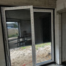 GPF FIRMA HANDLOWO USŁUGOWA - Zamknięte rolety zewnętrzne w oknie PCV Aluplast w surowym wnętrzu. Widoczne niedokończone ściany z betonu komórkowego i podłoże. Na zewnątrz drabina i trawa.