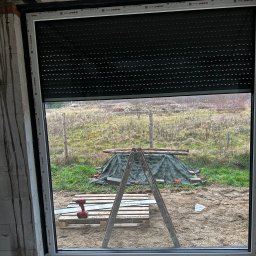 GPF FIRMA HANDLOWO USŁUGOWA - Montaż rolety zewnętrznej w oknie PCV na surowym etapie budowy. Widoczny wkrętak akumulatorowy, drabina i paleta. Za oknem widok na zewnątrz.