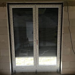 GPF FIRMA HANDLOWO USŁUGOWA - Białe okno PCV Aluplast z opuszczoną roletą zewnętrzną w surowym murze z betonu komórkowego. Widoczne mocowania okna. Częściowo widoczny widok za oknem.