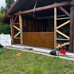 Montaż segmentowej bramy garażowej w drewnianej konstrukcji budynku gospodarczego, widoczne narzędzia i akcesoria montażowe, trawnik i częściowo ułożona kostka brukowa.