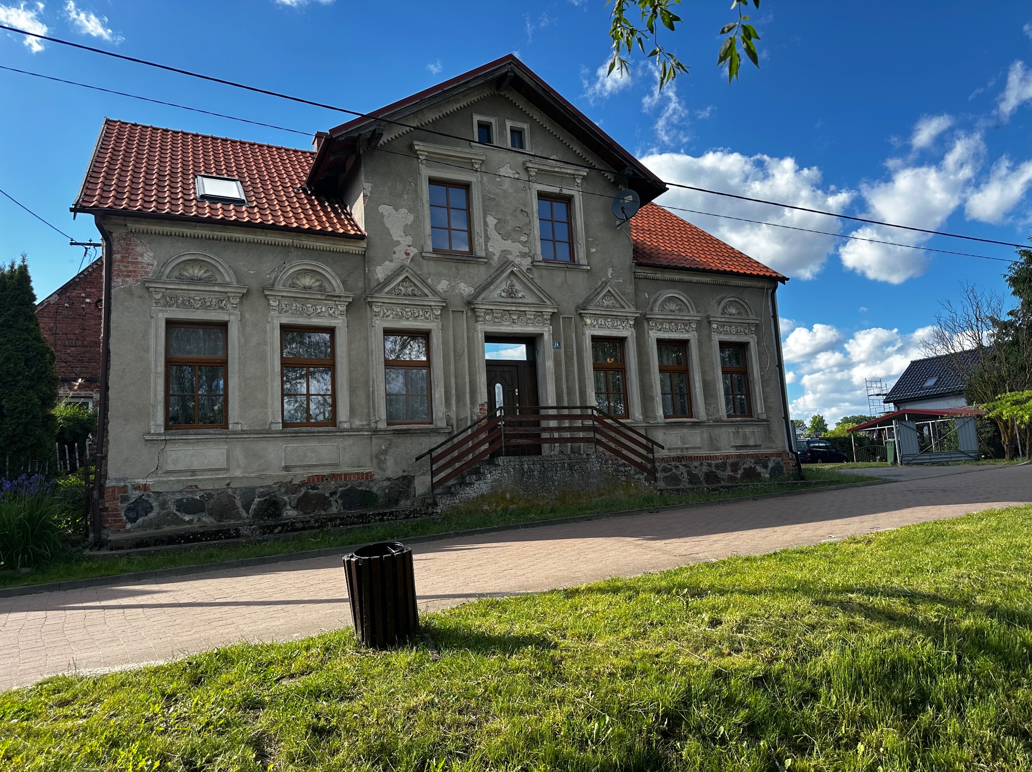 Od frontu szary, dwupiętrowy dom z czerwonym dachem, drewnianymi oknami i schodami wejściowymi. Na ścianie widoczna antena satelitarna. Błękitne niebo z chmurami w tle.