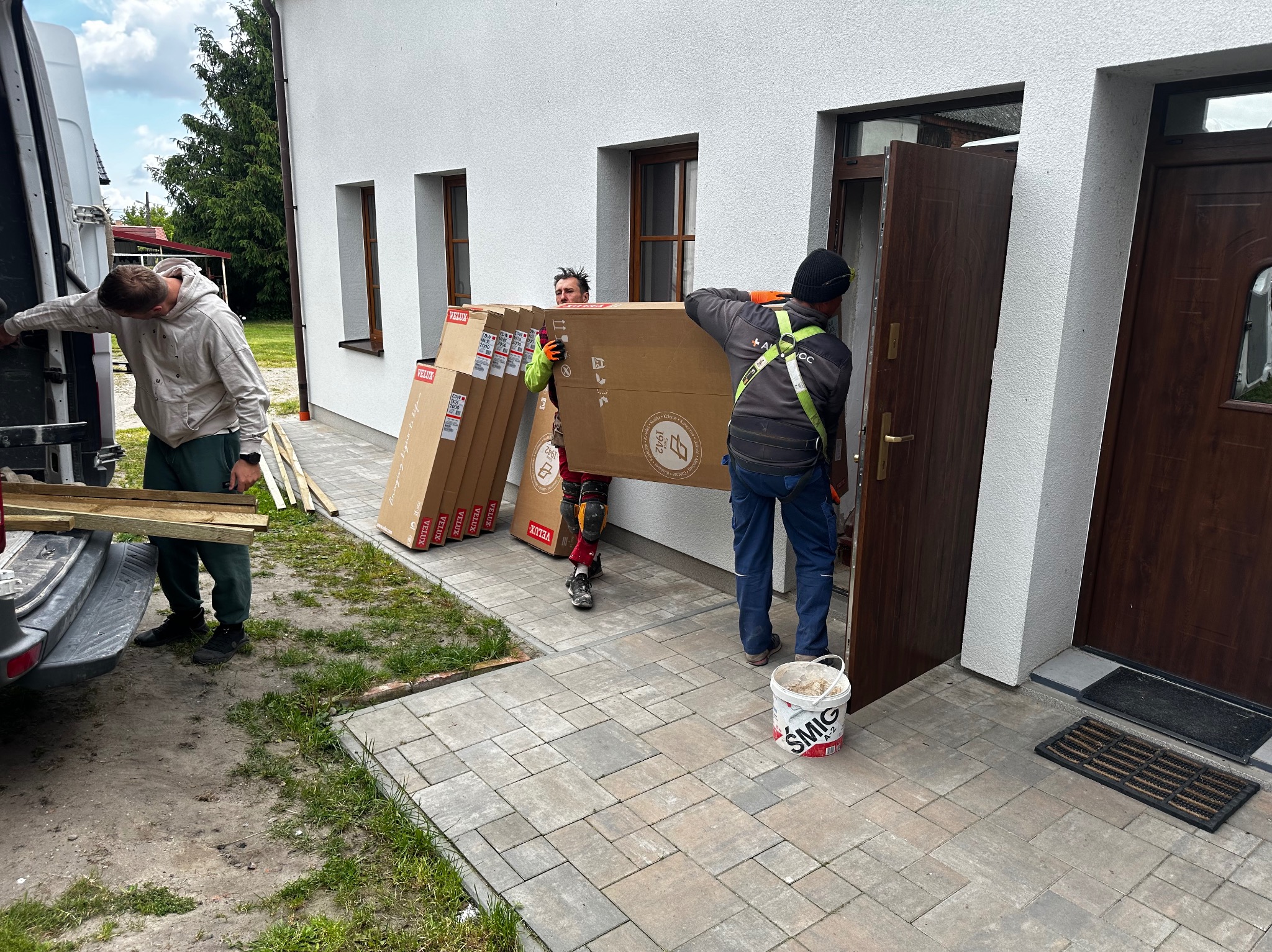 Montaż drzwi zewnętrznych: Dwóch monterów wnosi zapakowane drzwi do budynku. Widoczny bus dostawczy, narzędzia i wiadro z zaprawą. Dom z białej elewacji.