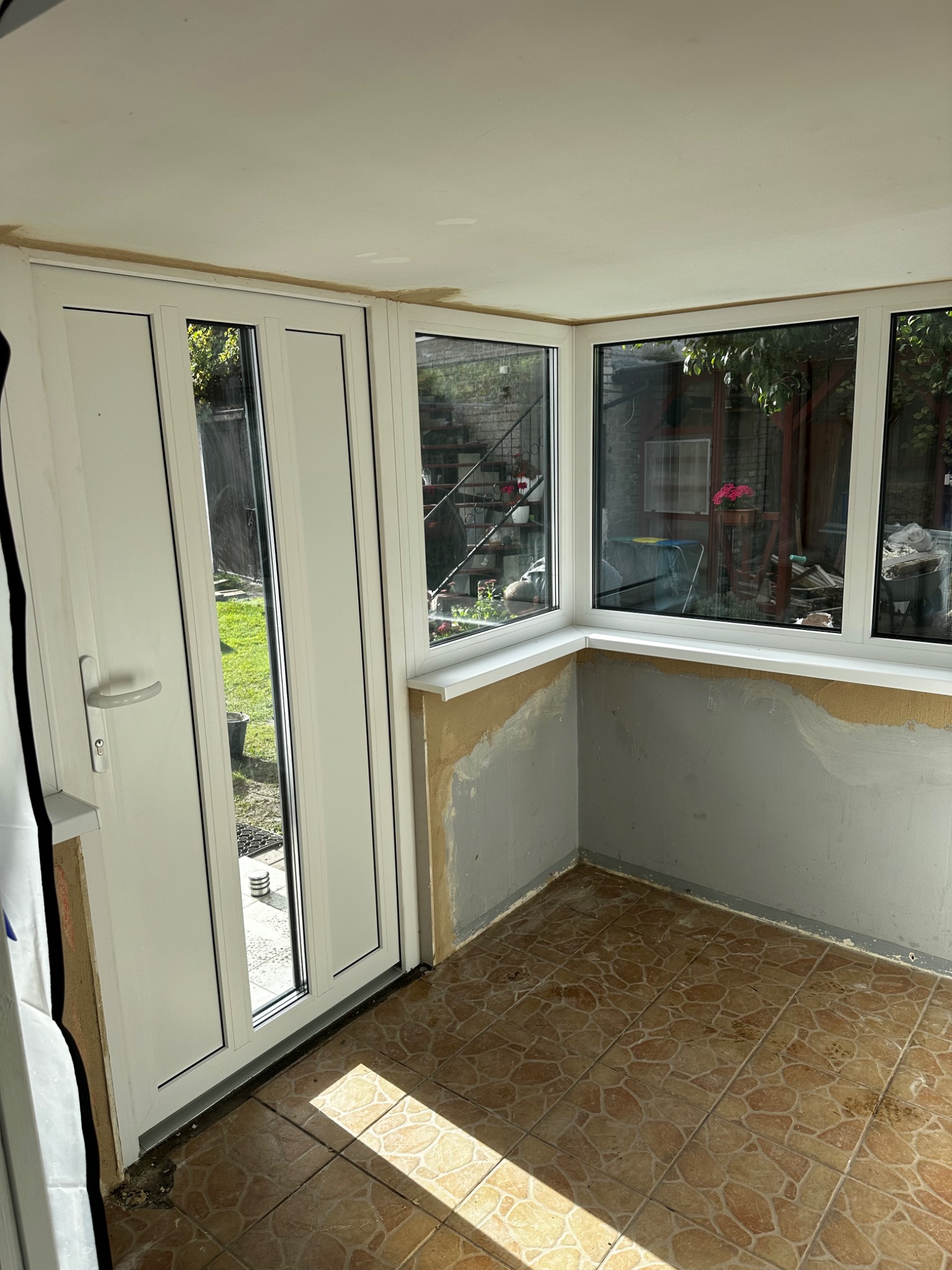 Jasne wnętrze z białymi oknami i drzwiami balkonowymi. Widoczne niedokończone ściany i brązowa podłoga. Za oknami zieleń ogrodu.