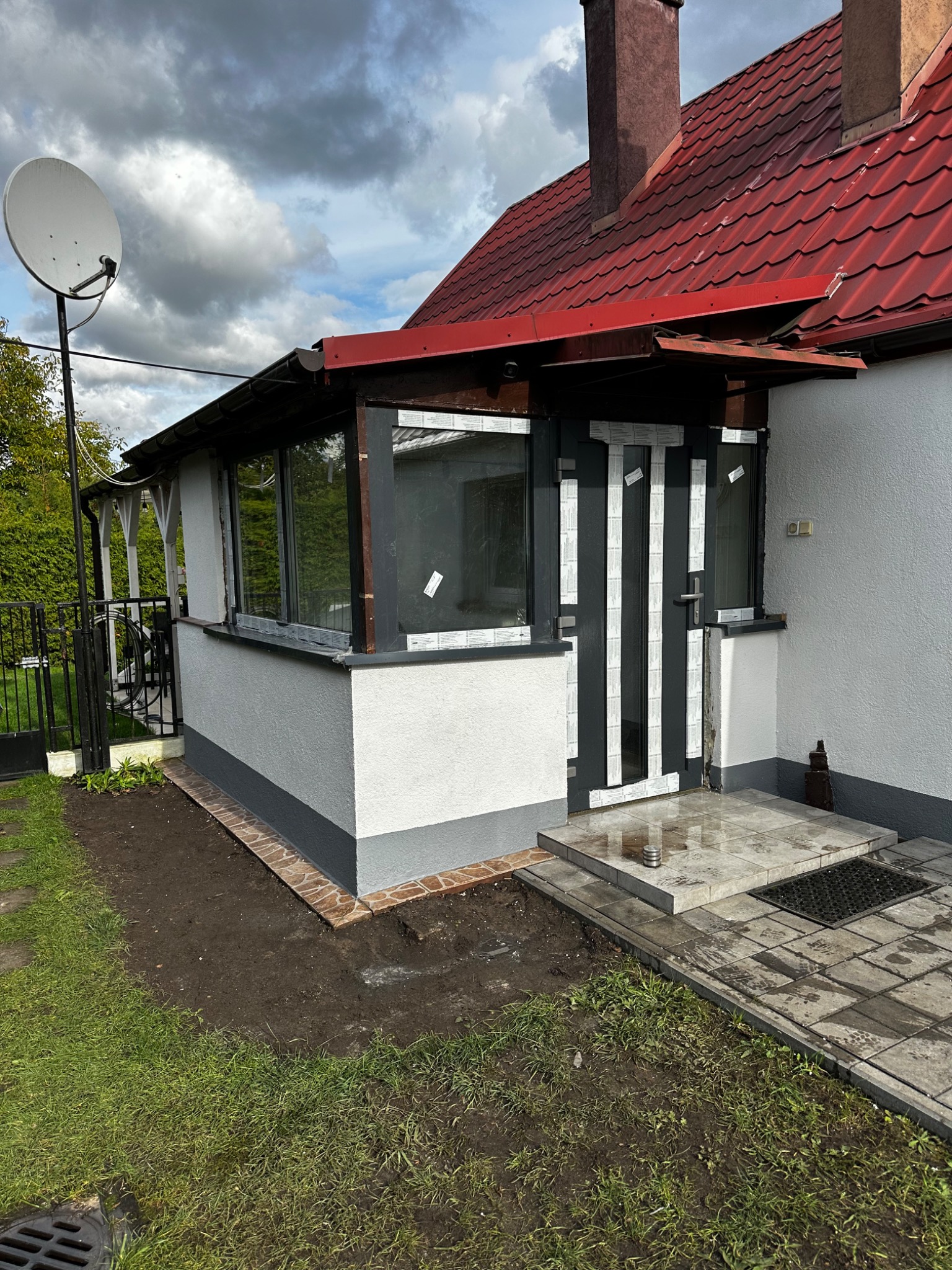 Zadaszone wejście do domu z oknami i drzwiami w trakcie montażu, zabezpieczone taśmą. Widoczny fragment elewacji, czerwony dach i trawnik. Ujęcie z poziomu gruntu.