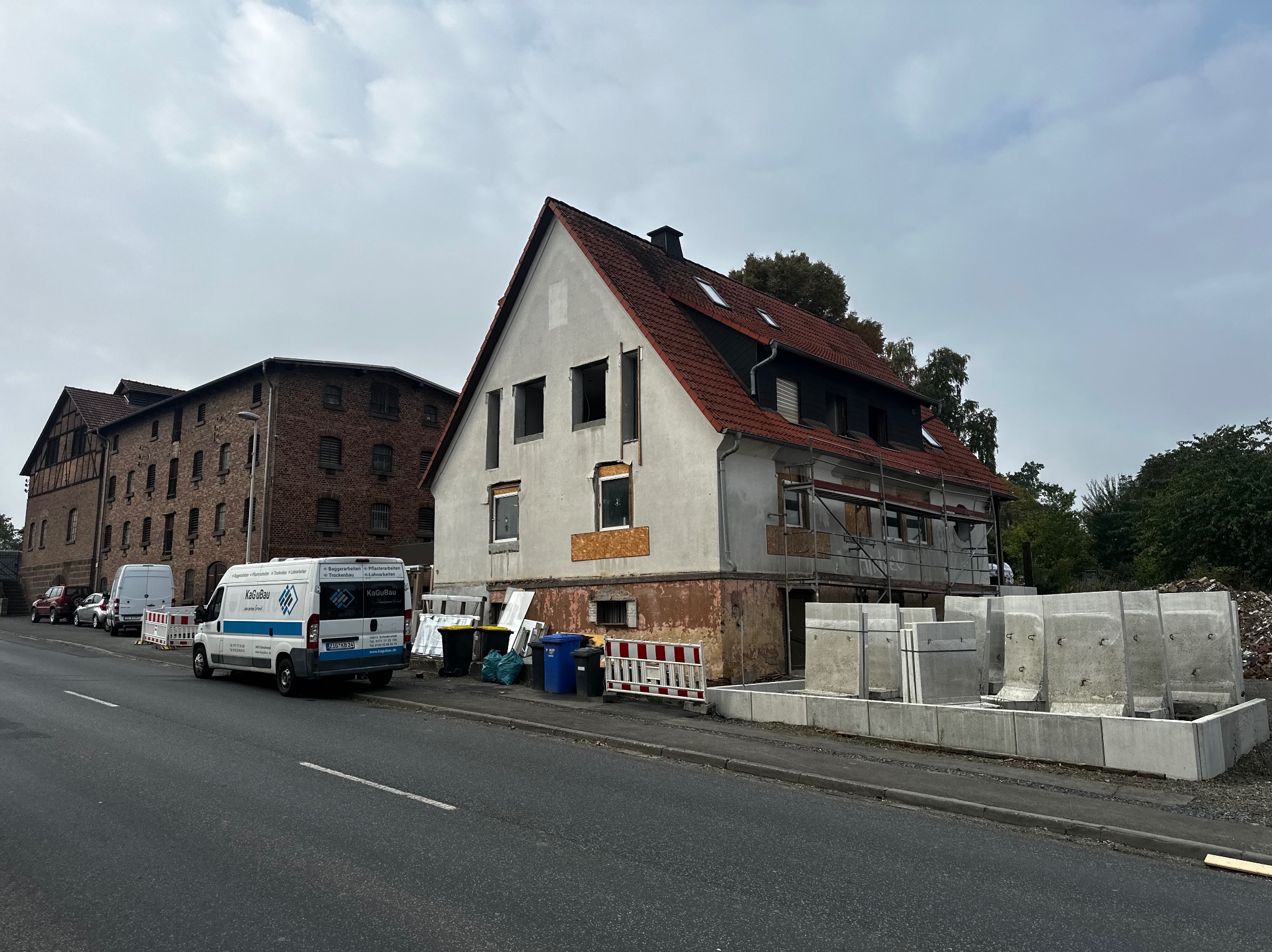 Dom w trakcie remontu z odsłoniętą elewacją, rusztowaniami i betonowymi elementami konstrukcyjnymi. Na pierwszym planie ulica i zaparkowany bus firmy budowlanej.