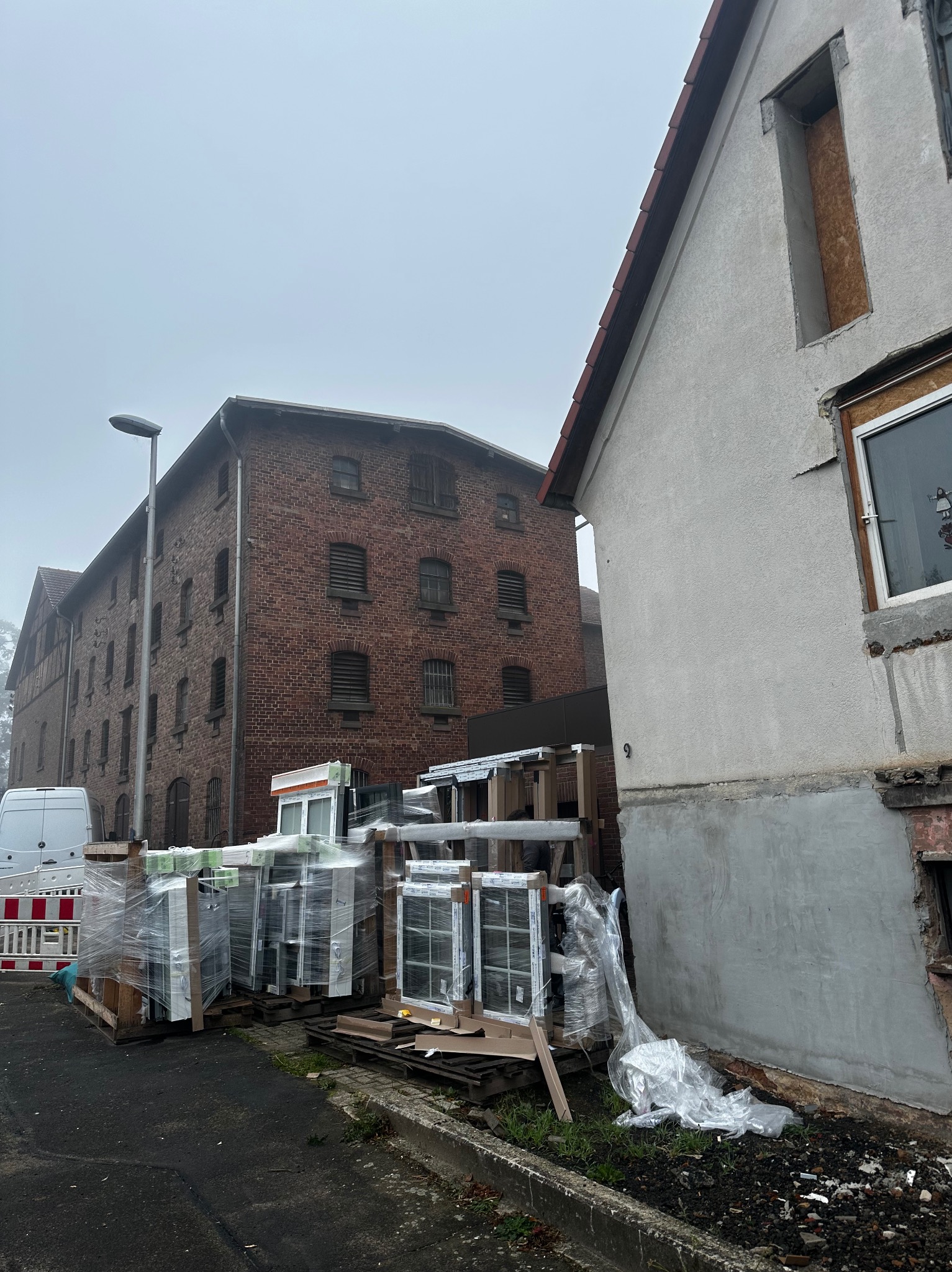 Pakiety okien PCV w folii na paletach przed budynkiem z cegły i otynkowanym domem w trakcie wymiany okien. Pochmurny dzień.