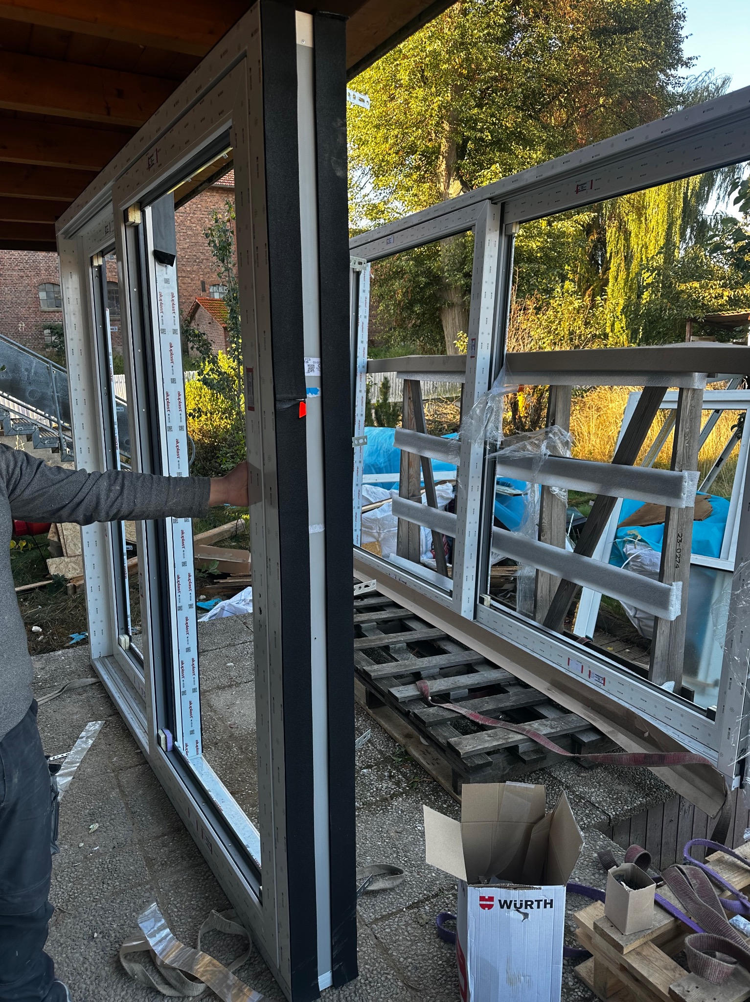 Montaż okna PCV: pracownik trzyma ramę okienną z czarną uszczelką, w tle inne ramy okienne zabezpieczone folią i karton Wurth na palecie. Widok z zewnątrz.