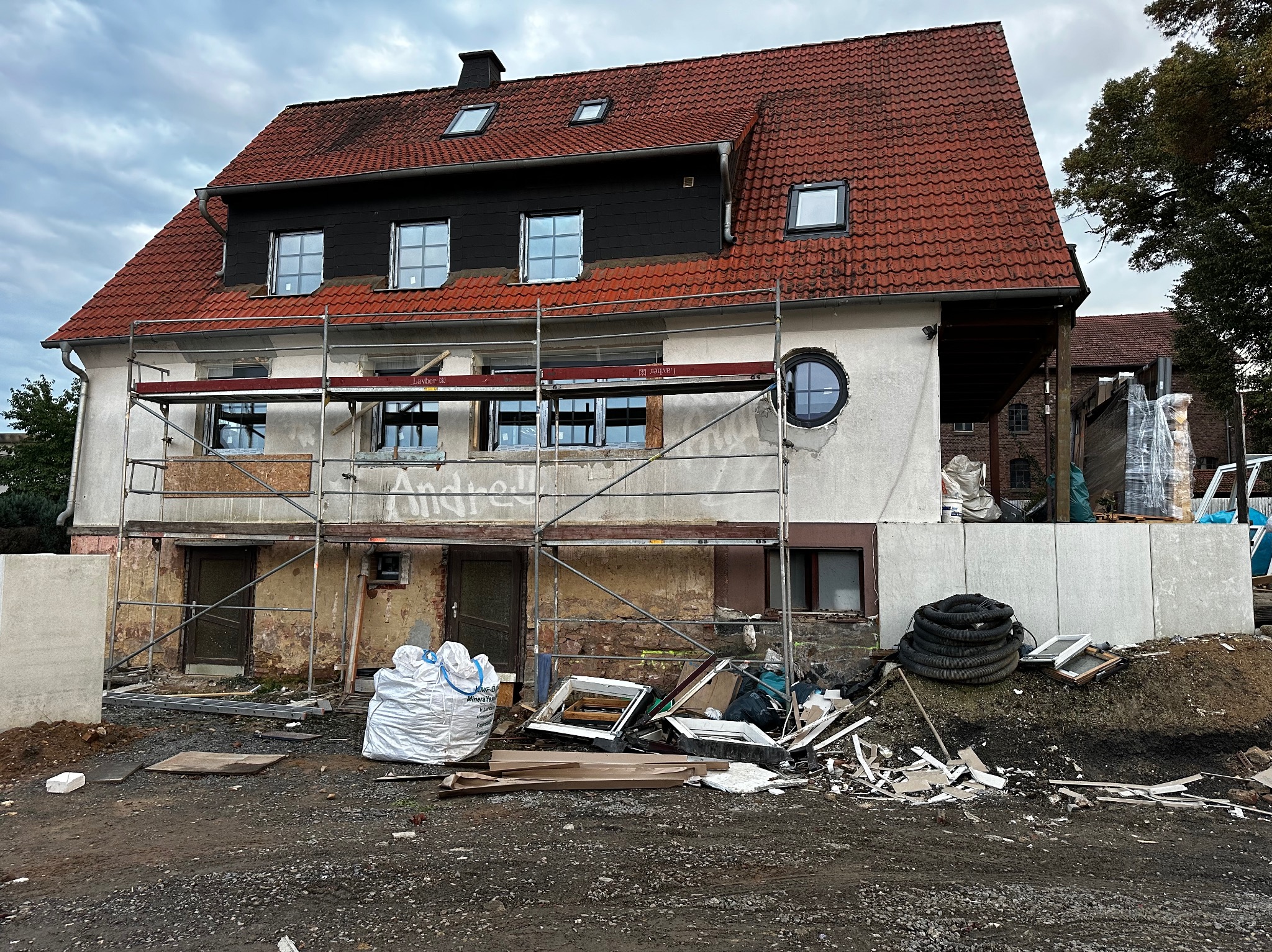 Dom w trakcie remontu elewacji z rusztowaniem. Widoczne stare okna, gruz i worki z materiałami budowlanymi. Częściowo odnowiona fasada i nowe okna na górze.