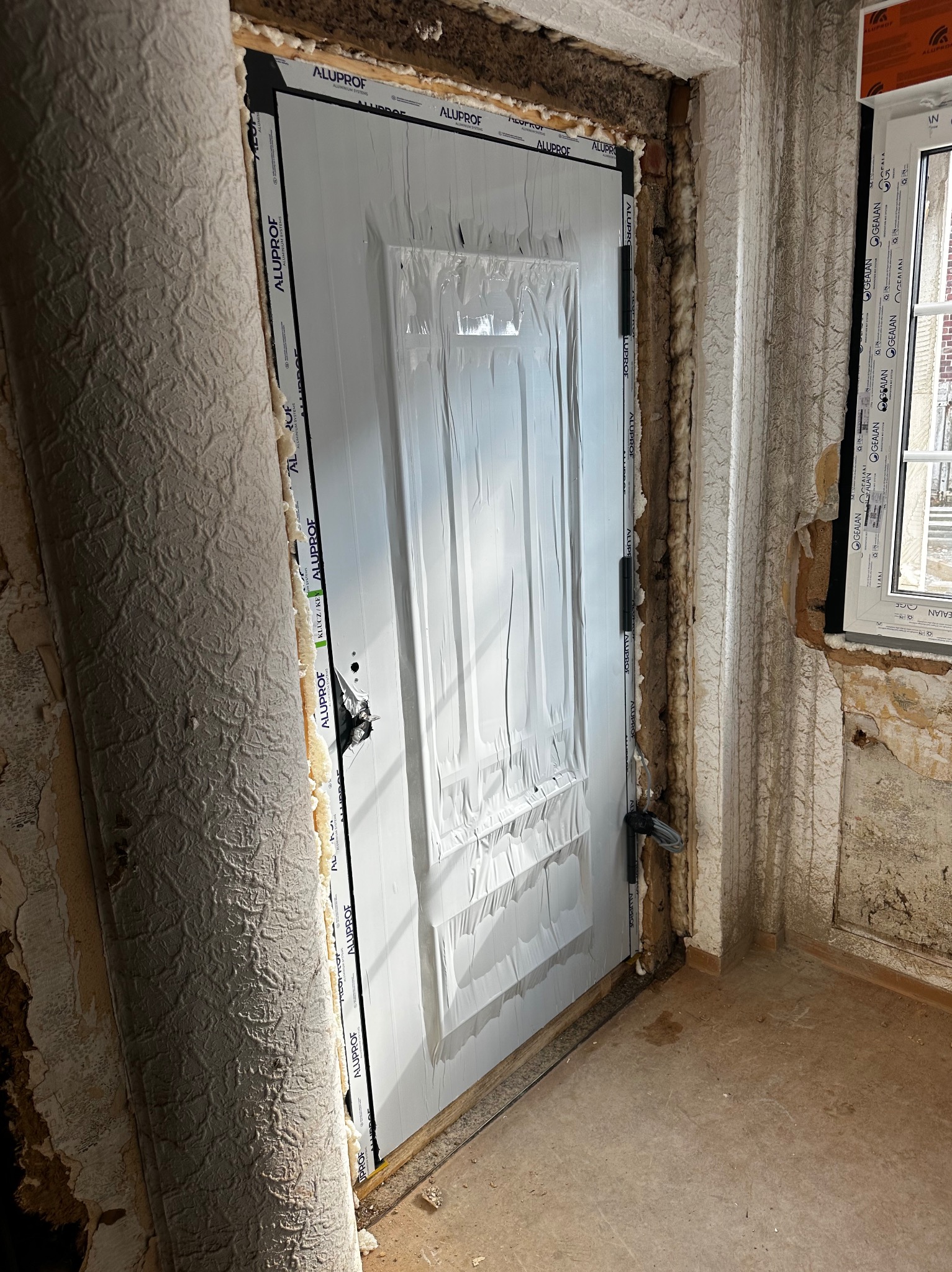 Montaż białych drzwi wewnętrznych Alufrof w surowym otworze, zabezpieczonych folią. Widoczne pianka montażowa i fragment okna Gealan. Ściany w trakcie remontu.