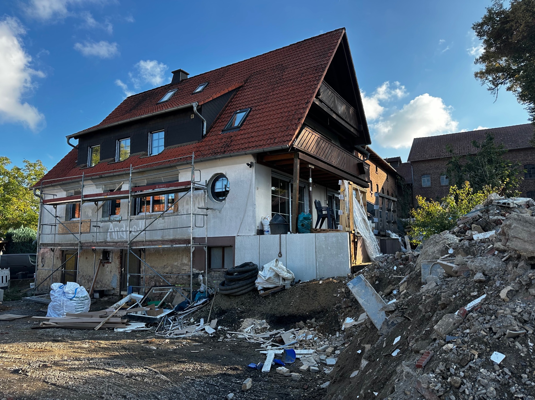 Dom w trakcie remontu: widoczne rusztowania, odsłonięte fragmenty elewacji, gruz. Częściowo odnowiona fasada z oknami i balkonem. Niebo z chmurami w tle.