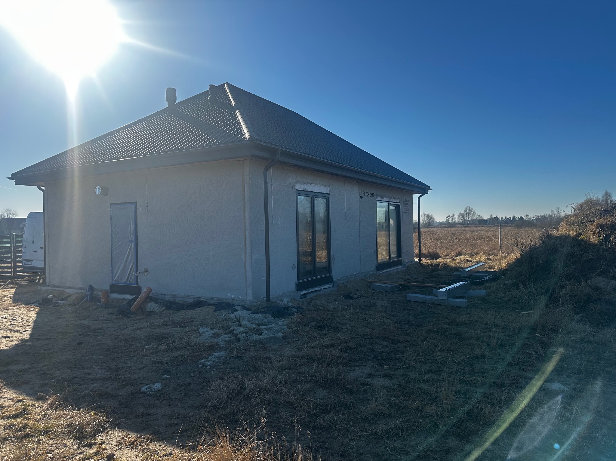 Nowoczesny dom jednorodzinny w trakcie wykończenia elewacji zewnętrznej, z widocznymi oknami i przygotowanym terenem wokół, w słoneczny dzień.