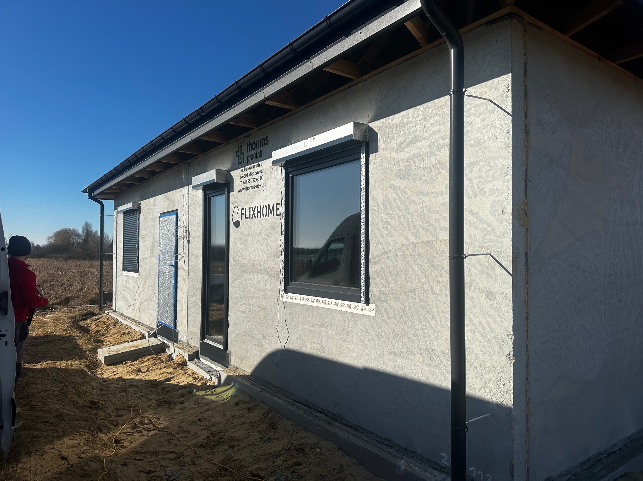 Dom w trakcie budowy z zamontowanymi oknami i roletami zewnętrznymi. Widoczne elementy konstrukcyjne i instalacyjne. Jasne tło nieba.