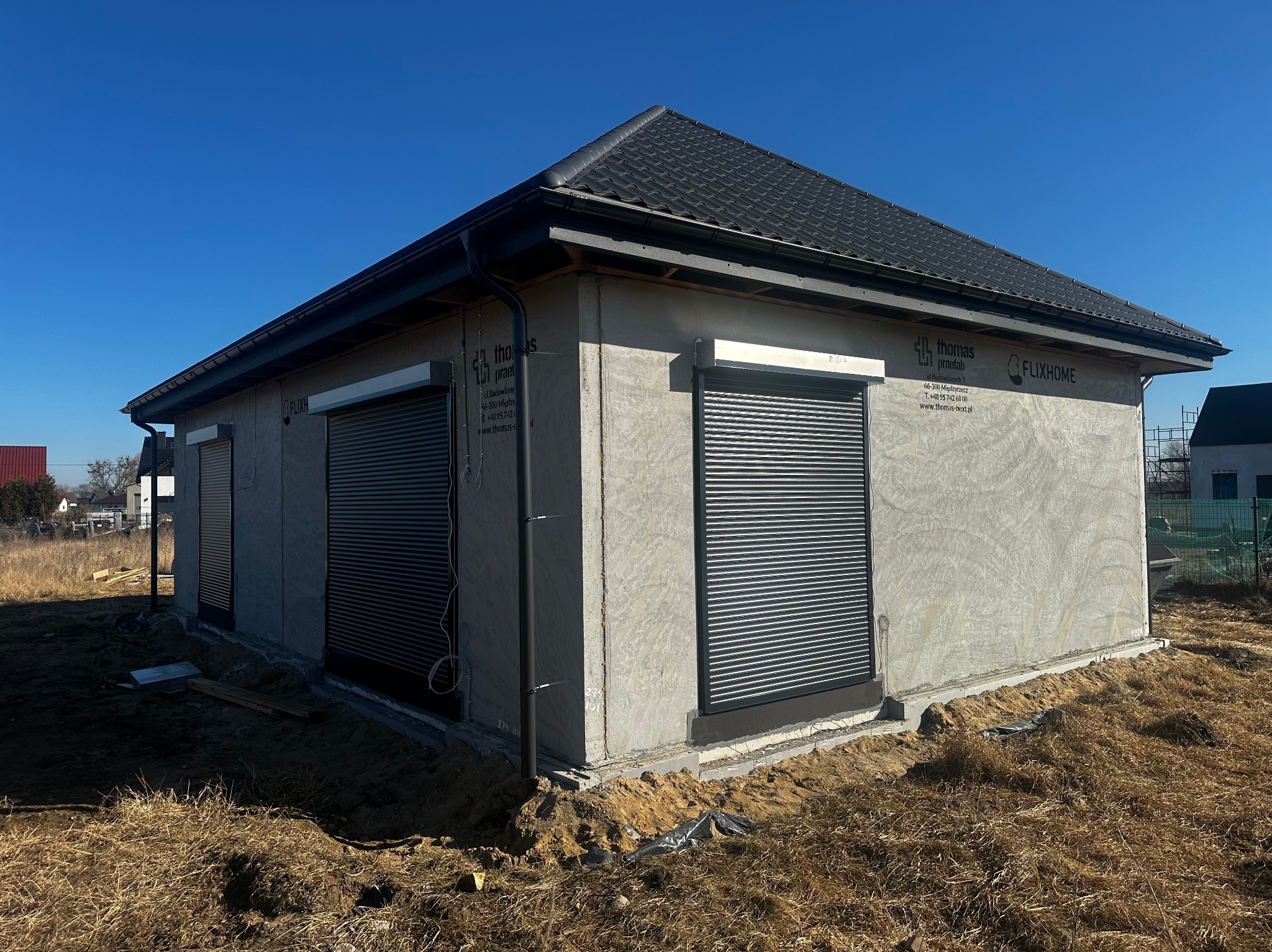 Dom w trakcie budowy z zamontowanymi roletami zewnętrznymi w kolorze szarym. Widoczne panele elewacyjne z logo producenta. Błękitne niebo w tle.