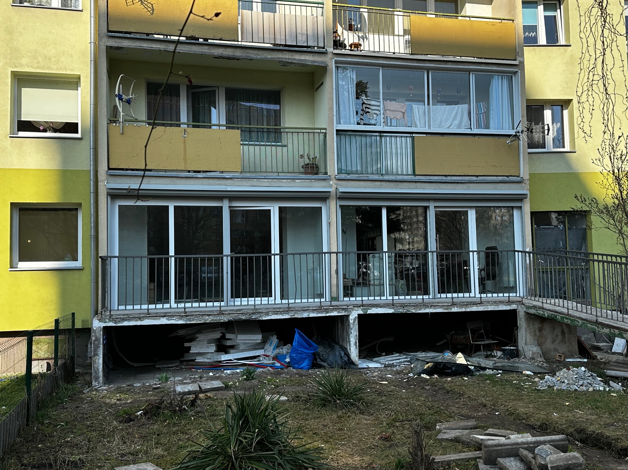 Dwukondygnacyjny budynek z balkonami i dużymi oknami, widoczne ślady prac remontowych na zewnątrz. Elementy budowlane porozrzucane wokół budynku.
