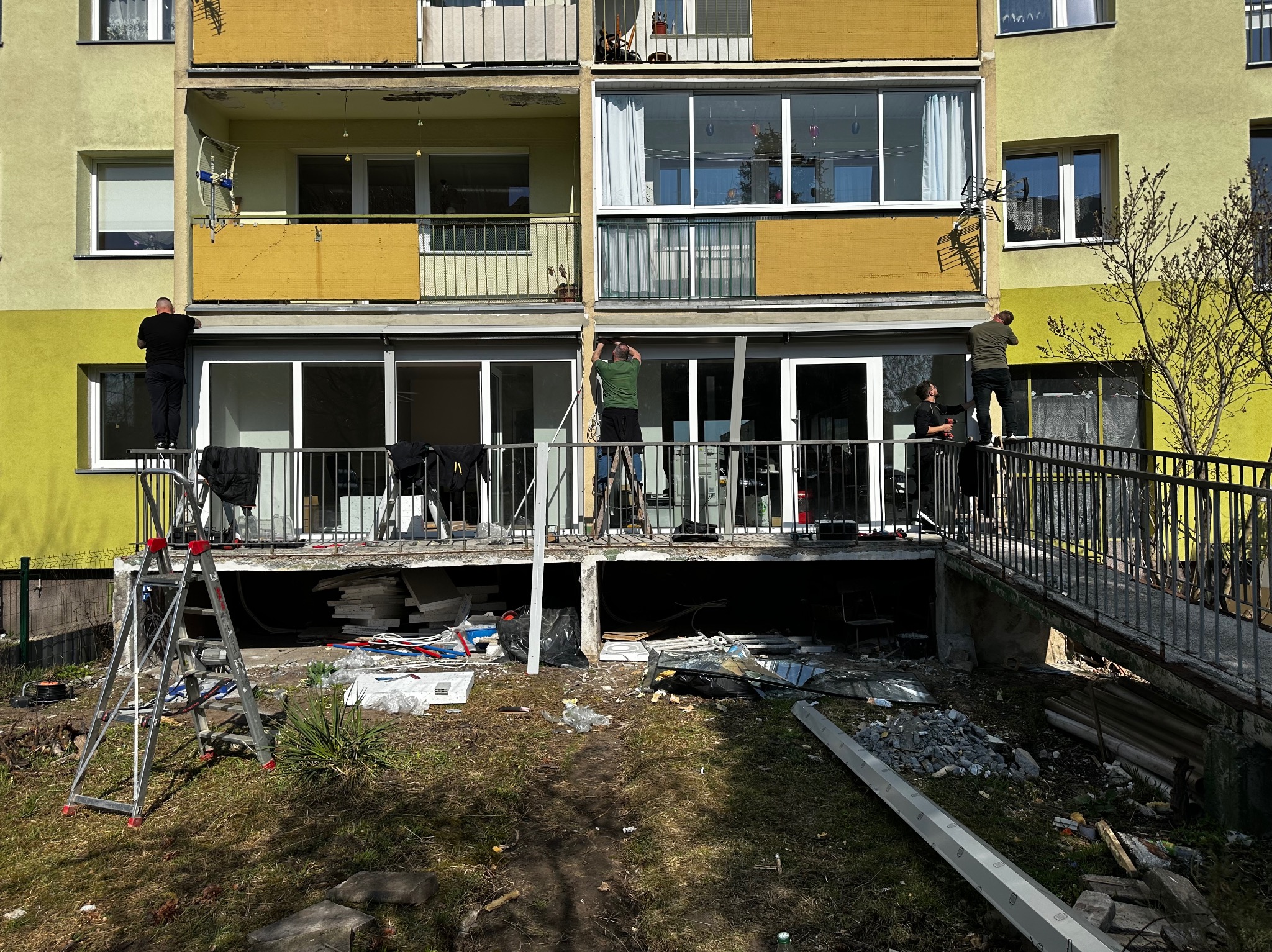 Montaż okien balkonowych w bloku z żółtą elewacją. Trzech monterów na drabinach i balkonie. Widoczne ślady demontażu i stare ramy okienne.