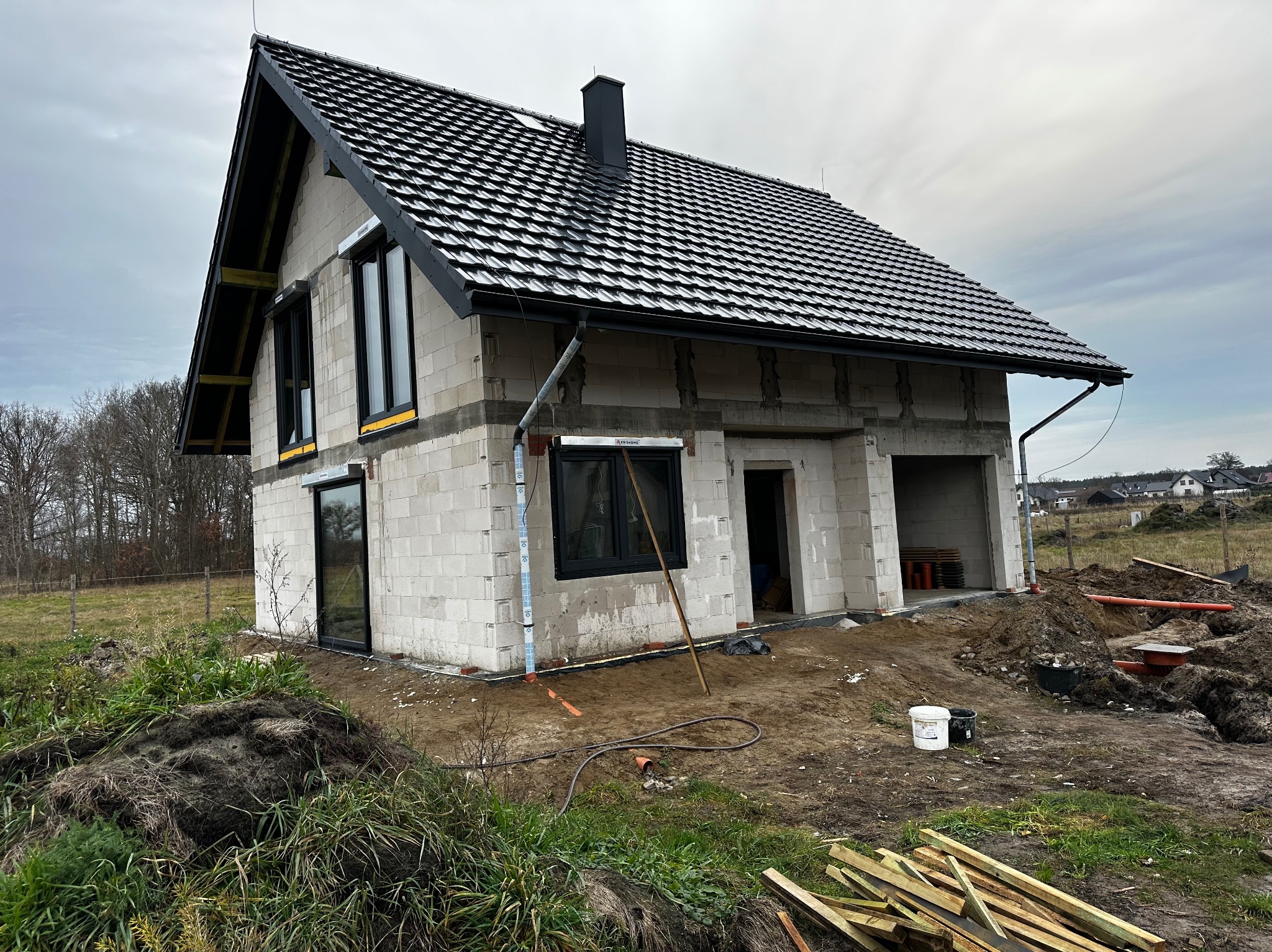 Surowy stan domu jednorodzinnego z czarną dachówką i oknami, widoczne bloczki betonowe, teren budowy z ziemią i drewnem, pochmurne niebo w tle.