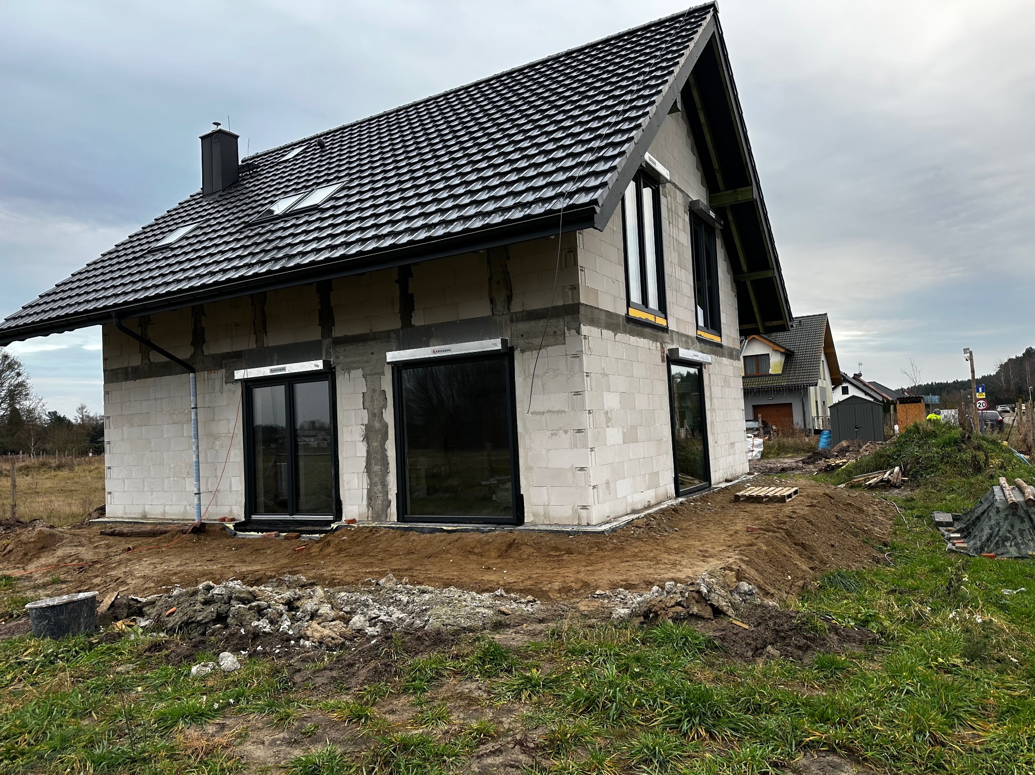 Surowy stan domu jednorodzinnego z czarnymi oknami i roletami zewnętrznymi w kolorze grafitowym. Widoczne bloczki betonowe i czarna dachówka.