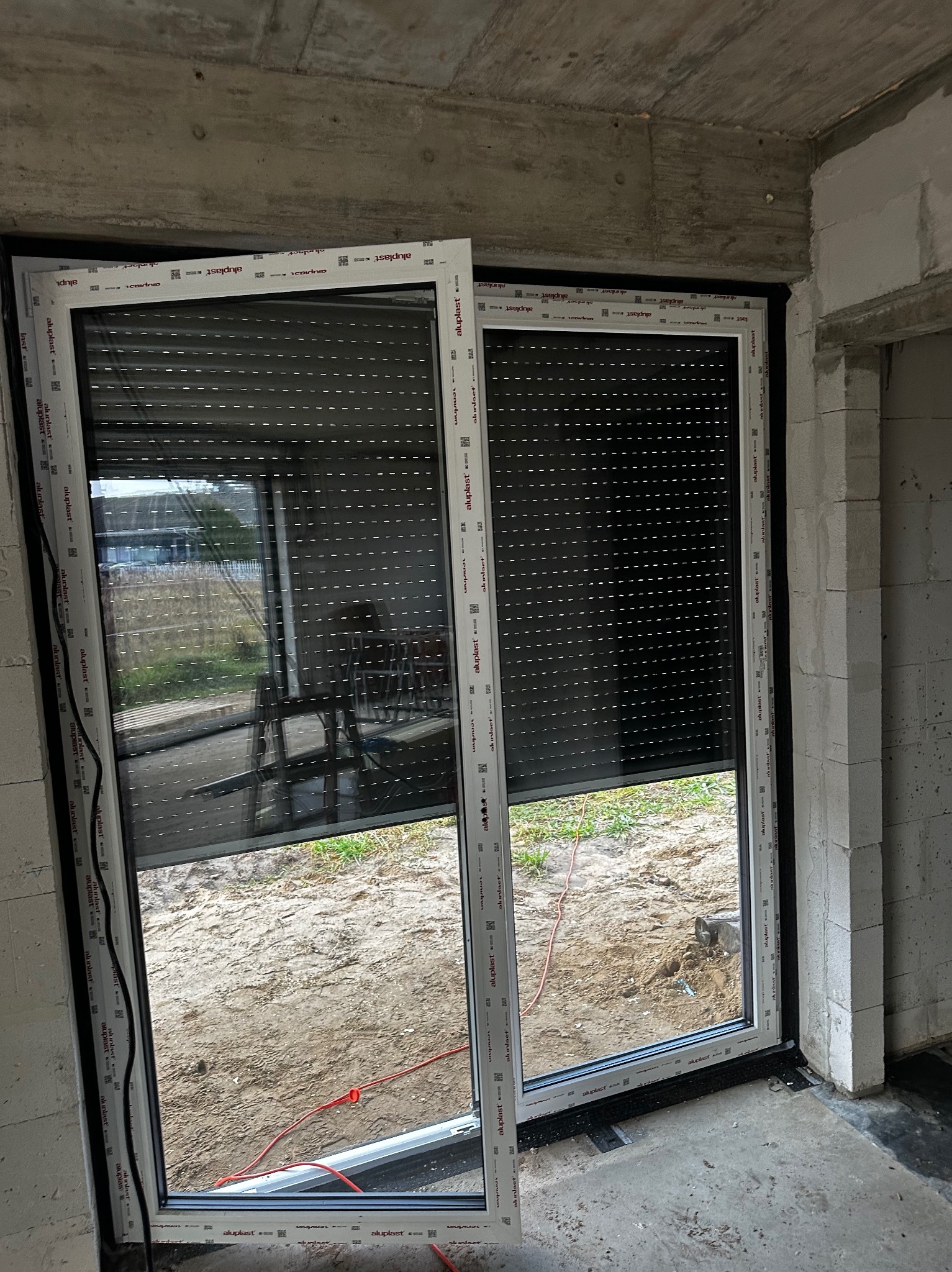 Zamknięte rolety zewnętrzne w oknie PCV Aluplast w surowym wnętrzu. Widoczne niedokończone ściany z betonu komórkowego i podłoże. Na zewnątrz drabina i trawa.