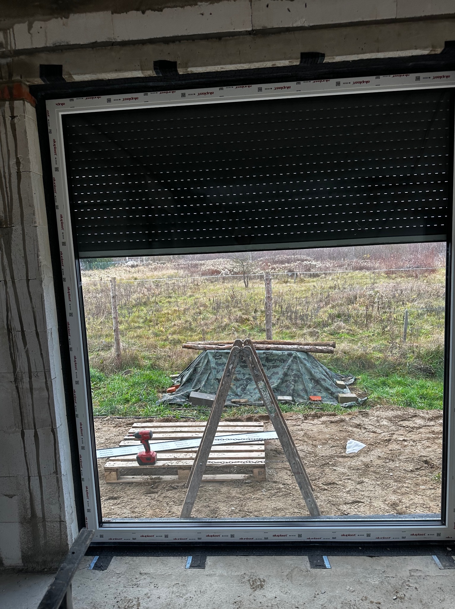 Montaż rolety zewnętrznej w oknie PCV na surowym etapie budowy. Widoczny wkrętak akumulatorowy, drabina i paleta. Za oknem widok na zewnątrz.